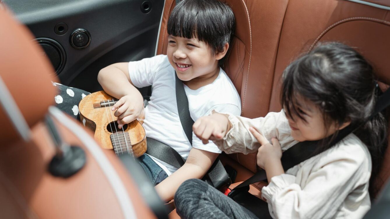 From above smiling ethnic boy and girl in casual outfits sitting fastened in passenger seats with ukulele during road trip together