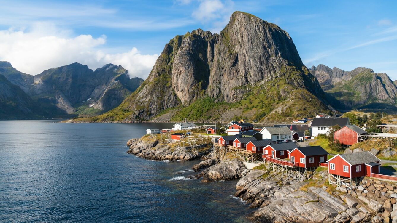 a group of houses by a body of water with mountains in the background with Lofoten in the background/ Photo by Benoît Deschasaux/Unsplash